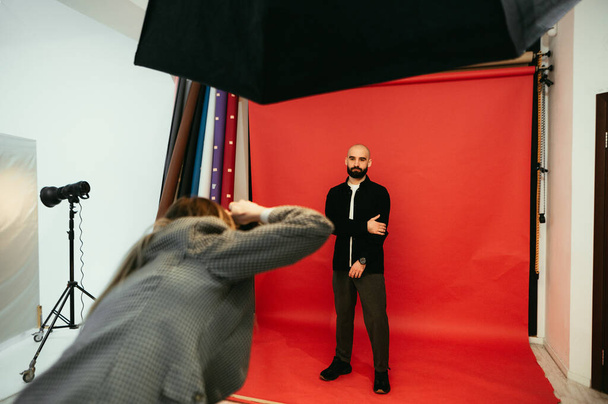 Fotógrafo hace una sesión de fotos para un hombre barbudo guapo sobre un fondo rojo en un estudio de fotografía - Foto, Imagen