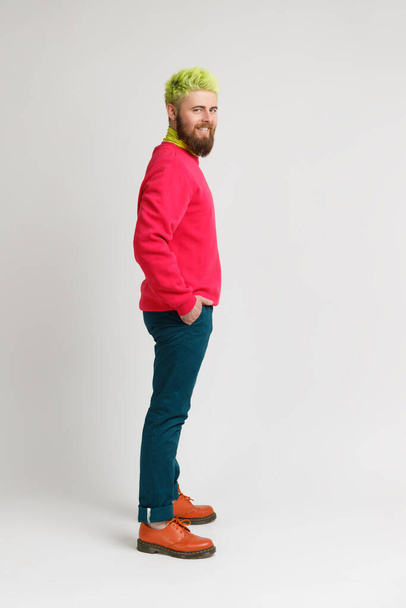 Vista lateral de hombre barbudo feliz expresa emociones positivas, mira a la cámara con sonrisa dentada, vistiendo suéter rojo y pantalones elegantes. Estudio interior plano aislado sobre fondo gris. - Foto, imagen