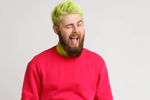 Cheerful playful young Caucasian man with beard in good mood, smiling broadly and winking at camera, keeps mouth open, dresses red jumper. Indoor studio shot isolated on gray background. - Foto, afbeelding