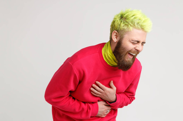Portrait of excited happy yellow-haired man with beard, dresses in casual red sweater, laughing loud, hearing really funny thing, keeps hands on belly. Indoor studio shot isolated on gray background. - Foto, immagini