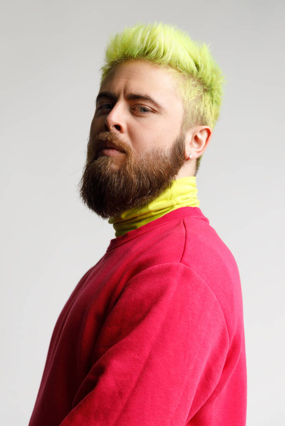 Portrait of proud handsome young bearded Caucasian man in red casual sweater, looking with prideful face,standing half sideways smug puffed. Indoor studio shot isolated on gray background. - Foto, Bild