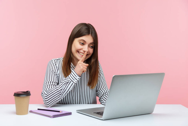 Please, keep secret. Positive woman in striped shirt shushing with silence gesture, talking on video call, having conference via laptop at home office. Indoor studio shot isolated on pink background - 写真・画像