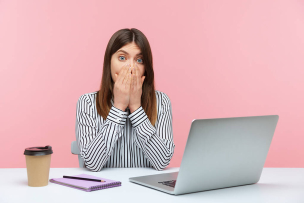 I will not tell anyone! Scared upset woman employee sitting at workplace with laptop and covering mouth with hand, afraid to tell corporate secret. Indoor studio shot isolated on pink background - Foto, Imagen