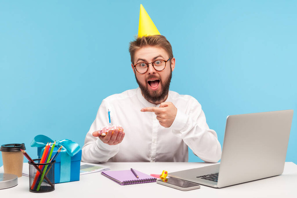 Funny excited man in eyeglasses and party hat pointing finger at little cupcake with candle, celebrating his company anniversary sitting at workplace. Indoor studio shot isolated on blue background - Photo, image