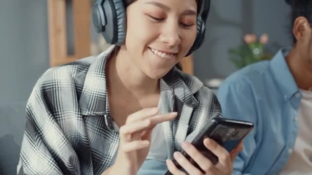 Šťastný mladý asijský pár aktivita muž použít notebook práce relaxovat si s ženami nosit sluchátka používat smartphone poslouchat hudbu na pohovce v obývacím pokoji v domě. Mladá vdaná práce z domova koncept. - Záběry, video