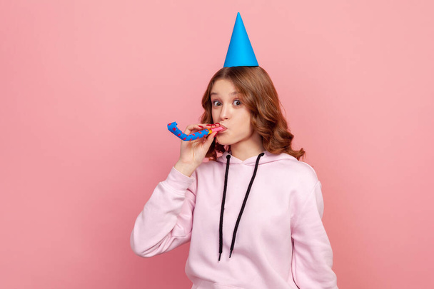 Portrait of happy curly haired teenage girl in hoodie with birthday hat blowing party horn, having fun at holiday celebration. Indoor studio shot isolated on pink background - Fotografie, Obrázek