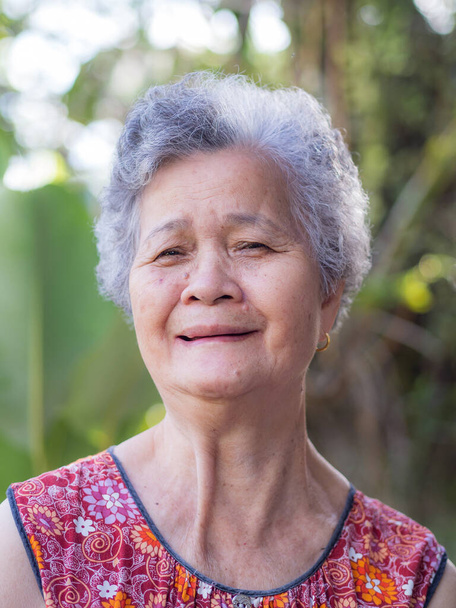 Vanhempi nainen, jolla on lyhyet valkoiset hiukset, seisoo hymyillen ja katsellen kameraa puutarhassa. Aasian vanhukset naiset terveitä ja on myönteisiä ajatuksia elämästä tehdä hänet onnelliseksi joka päivä. Terveysnäkökohdat - Valokuva, kuva