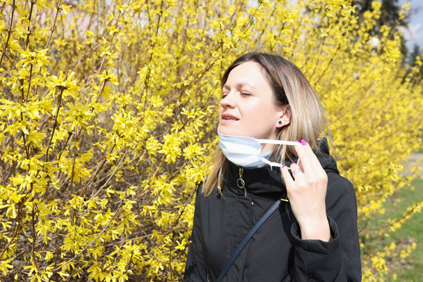 Femme aux yeux fermés enlevant le masque protecteur de son visage près d'un arbre en fleurs - Photo, image