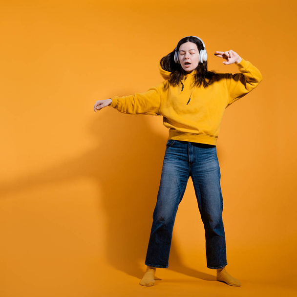 Happy dancing music lover. A young woman in a yellow hoodie with white wireless headphones, - Foto, Imagem