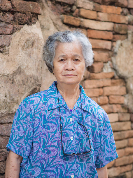 Портрет азіатської старшої жінки, яка дивиться на камеру, стоячи зі старим цегляним фоном. Концепція літніх людей і подорожі у відпустку. - Фото, зображення