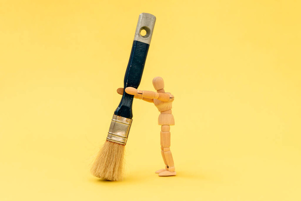 Wooden painter holding big paintbrush on the yellow background - Foto, afbeelding