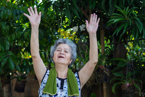 Portrait of an asian senior woman wearing casual clothing, smiling and exercises by gesturing raise both arms in her garden. Beautiful senior woman short hair with grey hair are happy and healthy - Photo, Image