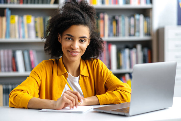 Portré aranyos sikeres afro-amerikai női diák ül asztalnál az egyetemi könyvtárban, visel stílusos ruhát, laptop, csinál házi feladatot, vagy felkészülés a vizsgára, nézi a kamera, mosolygós - Fotó, kép