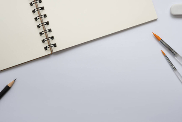 Cuaderno espiral abierto en blanco con pincel, lápiz y goma de borrar sobre fondo blanco, Espacio vacío para su texto, vista superior del espacio de trabajo. - Foto, Imagen