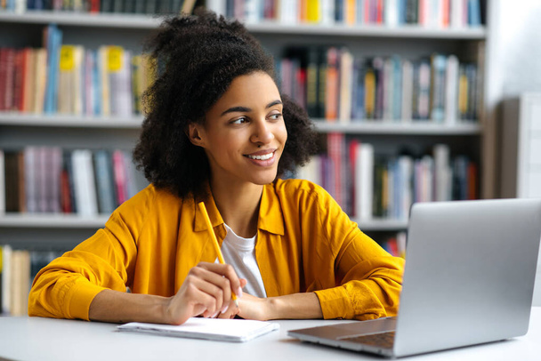 Χαρούμενη επιτυχημένη όμορφη σγουρά μαλλιά αφροαμερικανίδα φοιτήτρια κάθεται στο τραπέζι στη βιβλιοθήκη πανεπιστημιούπολη σε casual ρούχα, με φορητό υπολογιστή, κάνει την εργασία ή προετοιμασία για τις εξετάσεις, κοιτάζοντας μακριά, χαμογελώντας - Φωτογραφία, εικόνα