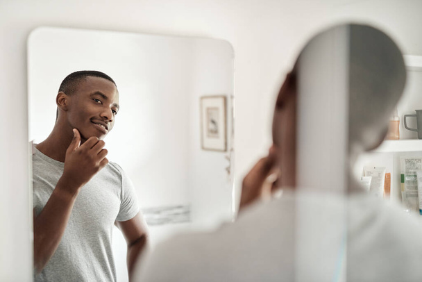 Улыбающийся молодой африканский мужчина трогает его лицо рукой в зеркале в ванной утром - Фото, изображение