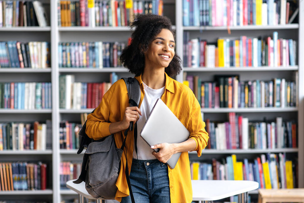 Portret van vrolijke mooie Afro-Amerikaanse studente, staande tegen de achtergrond van boekenplanken in de universiteitsbibliotheek met laptop en rugzak naar de zijkant kijkend, glimlachend - Foto, afbeelding