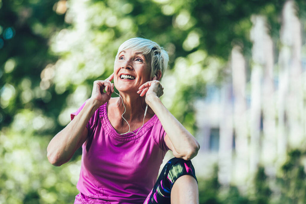 Mujer mayor haciendo ejercicio en el parque mientras escucha música. Mujer madura descansando después del ejercicio. - Foto, Imagen