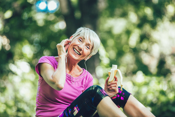 Senior woman exercising in park while listening to music. Mature woman eat banana resting after exercise. - Foto, afbeelding