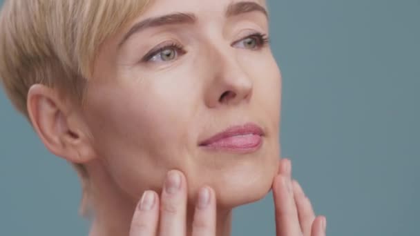 Cuidado de la piel antienvejecimiento. Primer plano retrato de la mujer madura haciendo masaje facial, piel de la cara hidratante, fondo azul - Metraje, vídeo