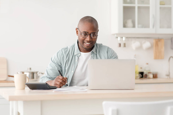 Planee las finanzas. Positivo afroamericano maduro hombre calcular los impuestos y el uso de ordenador portátil, sentado en la cocina - Foto, imagen