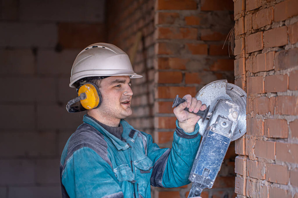Close-up de um construtor em um capacete de proteção em uma instalação de trabalho trabalha com uma ferramenta de corte. - Foto, Imagem