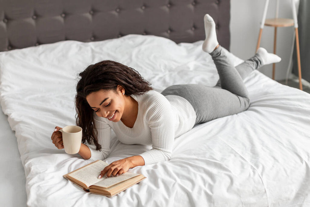 Schwarze Frau liest im Bett und hält Tasse heißen Kaffee in der Hand - Foto, Bild