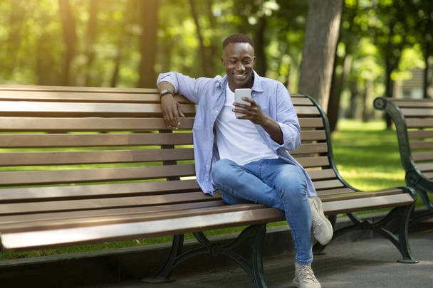 Mosolygó fekete fickó ül a padon a parkban, és használja az okostelefon, másolás helyet - Fotó, kép