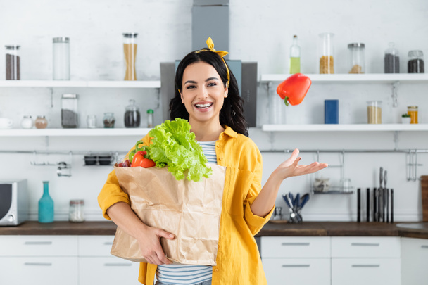 happy brunette woman throwing in air bell peppers and holding paper bag with groceries  - Foto, Bild