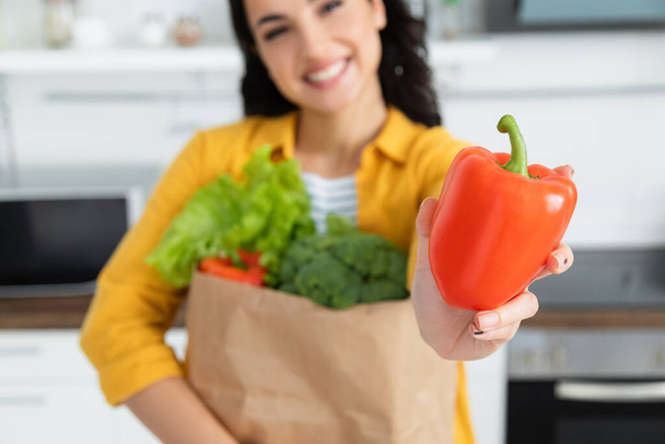 blurred and happy brunette woman holding paper bag with groceries and bell pepper  - Fotoğraf, Görsel