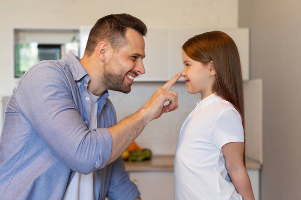 Papá alegre tocando la nariz de las pequeñas hijas posando en la cocina, con vista lateral - Foto, imagen