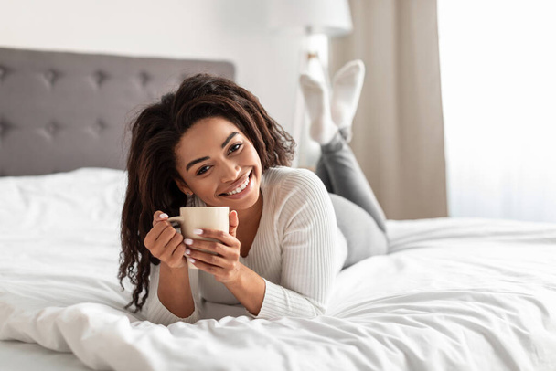Czarna kobieta leży na łóżku, trzymając filiżankę gorącej kawy - Zdjęcie, obraz