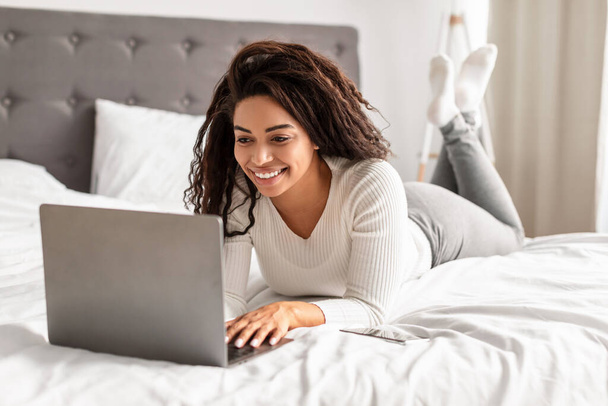 Szczęśliwa czarna kobieta leży w łóżku za pomocą laptopa - Zdjęcie, obraz
