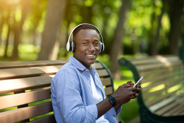 Handsome black man relaxing on bench in park and enjoying his favorite music on smartphone - Valokuva, kuva