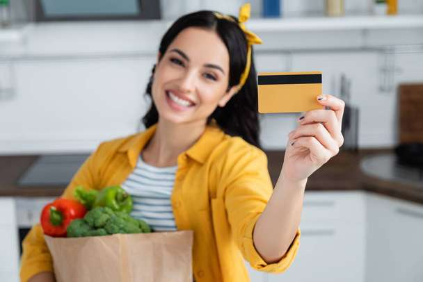 mujer feliz y borrosa sosteniendo bolsa de papel con comestibles y tarjeta de crédito - Foto, imagen