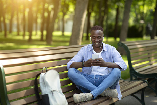 Funky black guy in earphones listening to his favorite music online from cellphone on bench at park, copy space - Фото, изображение
