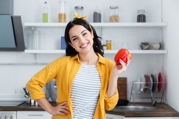 smiling brunette woman holding red bell pepper while standing with hand on hip - Фото, изображение