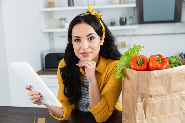 happy brunette woman using digital tablet near paper bag with fresh groceries  - Photo, Image