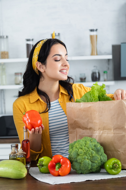 šťastná brunetka žena při pohledu na papírové tašky s čerstvými potravinami při držení papriky - Fotografie, Obrázek