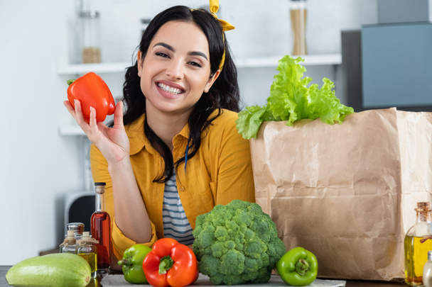 happy brunette woman smiling near paper bag with fresh groceries while holding bell pepper - Foto, Imagen