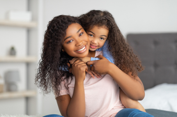 Láska mezi rodičem a dítětem. Pozitivní malé černošky dívka objímající její maminka na posteli doma - Fotografie, Obrázek