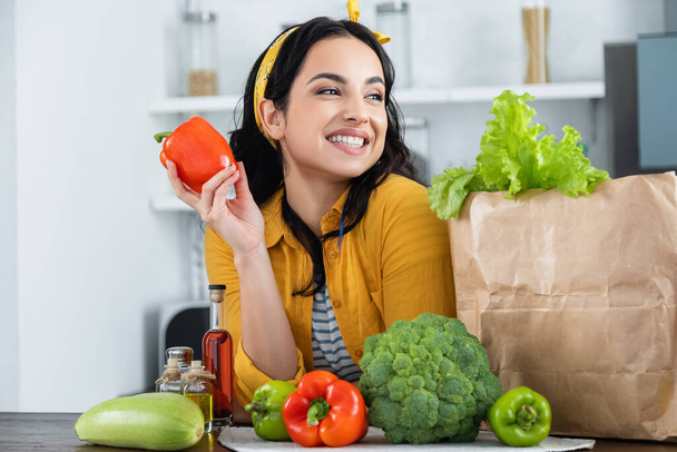 happy brunette woman smiling near paper bag with fresh groceries while holding red bell pepper - Photo, Image