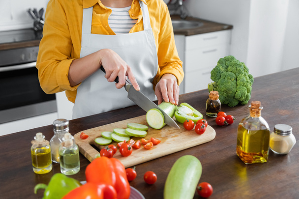 vista ritagliata di donna in grembiule taglio zucchine vicino pomodorini su tagliere  - Foto, immagini