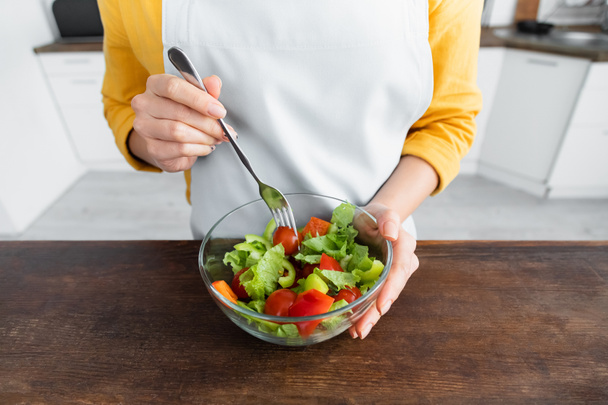 bijgesneden beeld van jonge vrouw in schort vork in de buurt van salade in kom - Foto, afbeelding