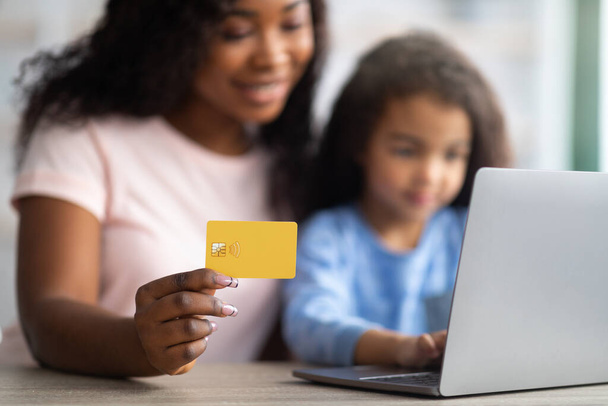 Compras online. Mãe afro-americana segurando cartão de crédito, fazendo compras na web com sua filha, usando laptop - Foto, Imagem