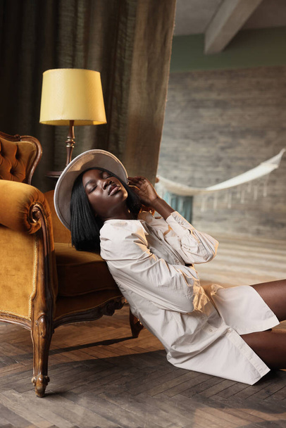 Photoshooting of a beautiful dark-skinned girl in the studio sitting on the floor near a chair, dressed in a beige dress and a hat. She poses in the camera. - Zdjęcie, obraz