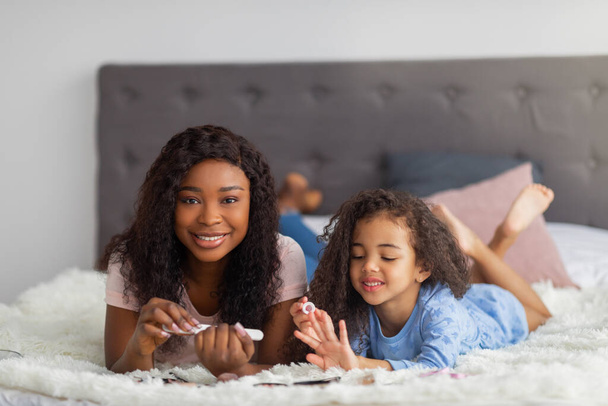 Fröhliche schwarze Mutter und ihre kleine Tochter bei Maniküre, zu Hause im Bett liegend - Foto, Bild
