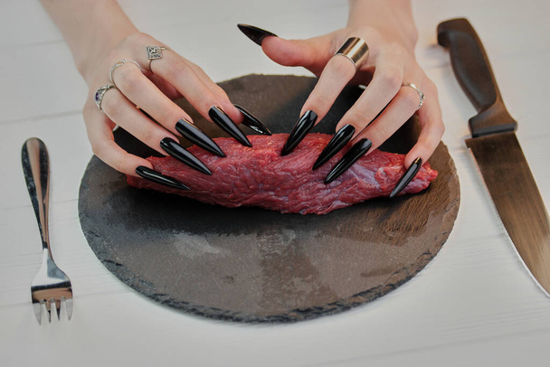 Weibliche Hände mit sehr langen schwarzen Nägeln, die rohes Steak berühren. - Foto, Bild