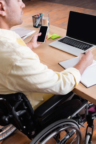 cropped view of disabled man holding smartphone with blank screen and writing in notebook - Photo, Image