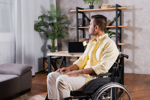 handicapped man sitting in wheelchair near laptop with blank screen on desk - Fotoğraf, Görsel